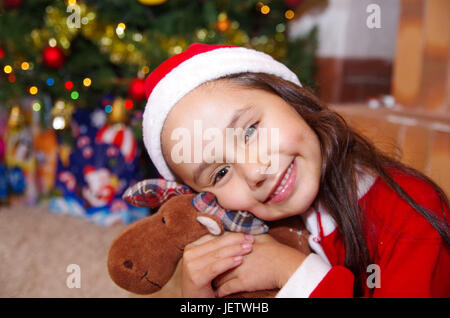 Schönes Lächeln auf den Lippen Litle Mädchen Kleidung ein Weihnachten, umarmt einen Elch Teddy mit einem Weihnachtsbaum-Hintergrund mit einigen Geschenken. Stockfoto