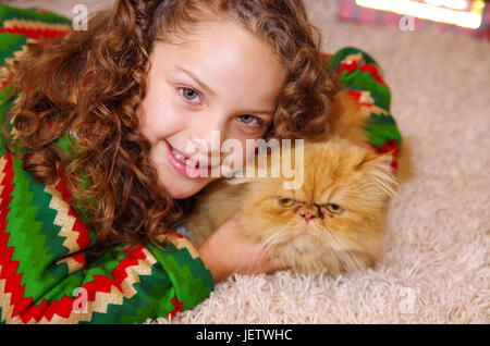 Schönes Lächeln auf den Lippen Litle Mädchen ein Weihnachten Kleidung, umarmt ihre gelbe Katze auf einem weißen Teppich verlegen. Stockfoto