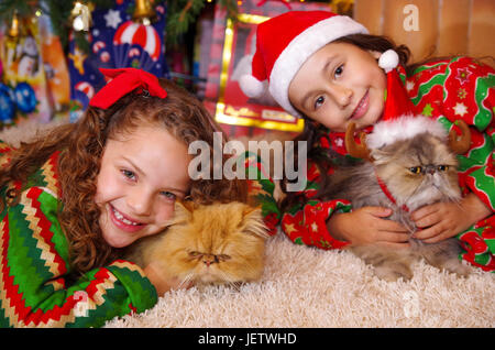 Schönes Lächeln auf den Lippen Litle Mädchen ein Weihnachten Kleidung, umarmt ihre gelbe Katze auf einem weißen Teppich verlegen. Stockfoto