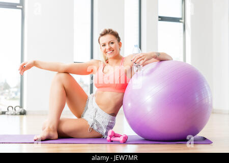Junge Frau entspannen nach Fitness-Workout. Fröhliche Sportlerin sitzt neben einem Fitball. Gesunde Lebensweise. Stockfoto
