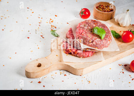 Frische rohe selbstgemachte Hackfleisch Steak Burger mit Gewürzen, Tomaten und Basilikum, auf einem weißen Stein Beton Tisch, Textfreiraum Stockfoto