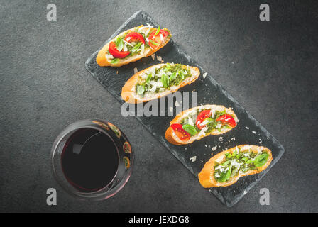 Italienisches Essen. Snacks. Antipasti. Bruschetta mit Pesto-Soße, Parmesan-Käse, Tomaten und frischem Basilikum. Auf schwarzen Tisch auf Schiefer. Mit Glas Stockfoto