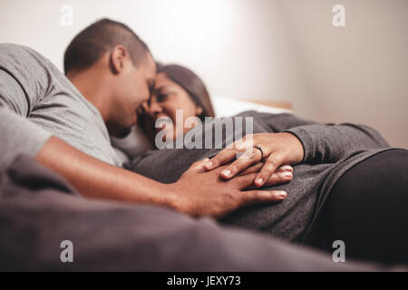 Romantischer junger Mann und Frau auf Bett, Fokus auf Hände auf Bauch liegend. African schwanger paar erwartet Baby. Stockfoto