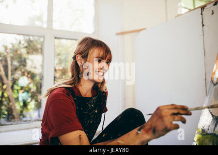 Innenaufnahme der Künstlerin Malerei auf Leinwand im Atelier und lächelnd. Glückliche junge Frau Maler Malerei in ihrer Werkstatt. Stockfoto
