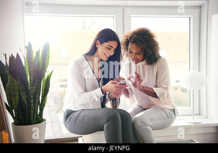 Zwei junge multirassische Unternehmerinnen auf Fensterbank am Arbeitsplatz sitzen und Surfen im Internet mit dem Smartphone. Stockfoto