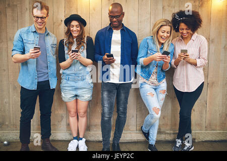 Schwarze und weiße Männer und Frauen stehen und SMS mit Smartphones bei Holzwand lächelnd. Stockfoto