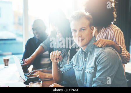Gut aussehender Mann hält Brillen sitzen am Konferenztisch mit Kollegen. Helle Fenster im Hintergrund. Stockfoto