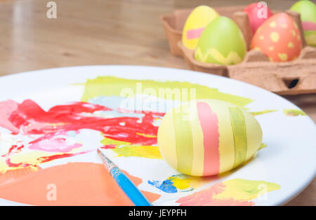 Eine gemütliche Osterei-Malerei-Szene auf einem Holztisch.  Fertige Eiern in einem Karton, Ei-in-Progress, Farben und Pinsel auf einer China-Teller-Palette in Stockfoto