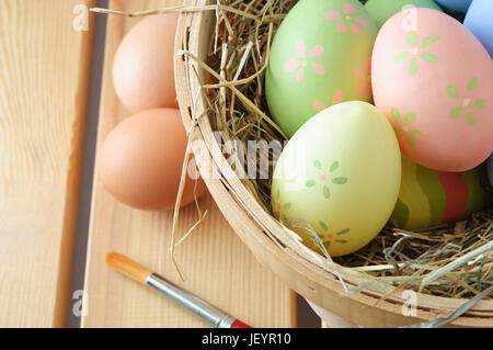 Nahaufnahme von Hand bemalte Ostereier in einem Korb, gefüllt mit Stroh mit natürlichen, unbemalt, Eiern und Pinsel auf ein Holzbrett Tischplatte ruht. Stockfoto