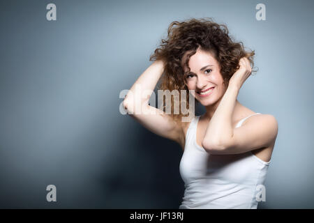 Junge Frau hält ihr lockiges Haar. Schön, lächelnd, positive Mädchen. Casual Portrait auf grauem Hintergrund. Natürliche Schönheit und Hapiness Konzept. Stockfoto