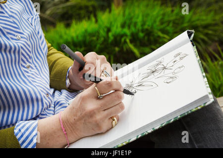 Nahaufnahme einer Frau sitzt in einem Garten, Blumen in einem Skizzenbuch zeichnen. Stockfoto