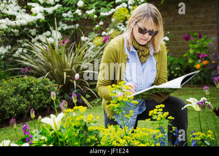 Frau mit Sonnenbrille steht in einem Garten durch ein Blumenbeet, in einem Skizzenbuch zeichnen. Stockfoto