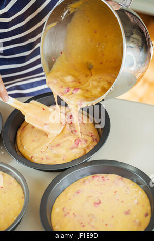 Nahaufnahme hohen Winkel der Person trägt ein blau-weiß gestreifte Schürze strömenden Kuchenteig aus einer Schüssel in runden Backformen Metall. Stockfoto