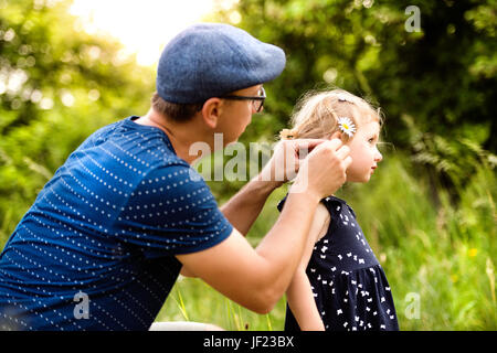 Junger Vater mit seiner niedlichen kleinen Tochter Zeit miteinander zu verbringen außerhalb im grünen Sommer Natur ihre Frisur zu tun. Stockfoto