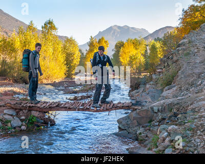 2 junge Männer trekking in der M'Goun Region des marokkanischen Atlas über einen Bach auf einer kleinen Holzbrücke Stockfoto