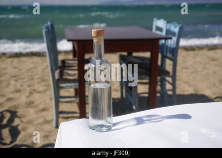Glasflasche Ouzo, Tsipouro Getränk von griechischen Strand mit unscharfen Taverne Tisch und Holzstühle blauen Hintergrund. Stockfoto