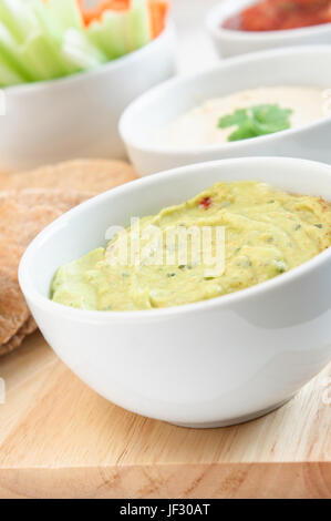 Guacamole, Hummus und Salsa dips in weißen Schalen auf einem Holzbrett mit Vollkorn Fladenbrot und Crudites beschnitten im linken Frame. Stockfoto