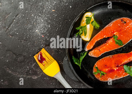 Frischen rohen unvorbereitet Fisch Lachs oder Forelle, Steaks in der Pfanne garen, mit Salz, Pfeffer, Zitrone und Barbecue sauce zum Grillen. Auf schwarzem Stein Stockfoto