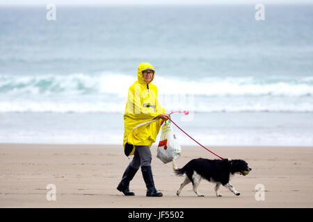 Eine Person zu Fuß einen Hund an der Leine entlang der unberührten und Hund freundlich Strand während einer Sommerdusche trägt einen gelben Regenmantel an Freathy an der Whitsand Bay, Cornwall, England Stockfoto