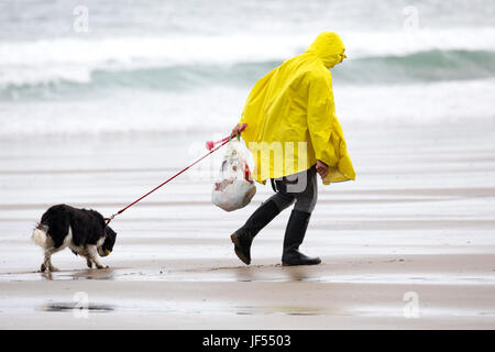 Eine Person zu Fuß einen Hund an der Leine entlang der unberührten und Hund freundlich Strand während einer Sommerdusche trägt einen gelben Regenmantel an Freathy an der Whitsand Bay, Cornwall, England Stockfoto