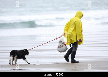 Eine Person zu Fuß einen Hund an der Leine entlang der unberührten und Hund freundlich Strand während einer Sommerdusche trägt einen gelben Regenmantel an Freathy an der Whitsand Bay, Cornwall, England Stockfoto