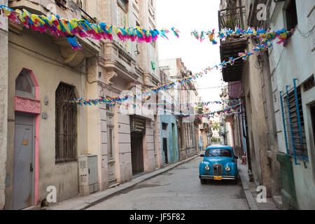 Oberhalb eines amerikanischen Oldtimers hängen bunte Dekorationen von Gebäuden auf einer schmalen Straße im Zentrum von Havanna. Stockfoto