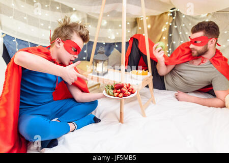 Glücklicher Vater und Sohn in Superhero Kostüme Früchte in Decke fort Stockfoto