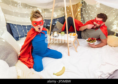 Glücklicher Vater und Sohn in Superhero Kostüme Früchte in Decke fort Stockfoto