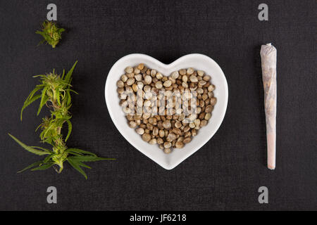 Gesunden Marihuana Medizin. Knospen, Samen in Herzförmige Schüssel und Cannabis gemeinsam von oben auf schwarzem Hintergrund. Ich liebe Alternativmedizin. Stockfoto