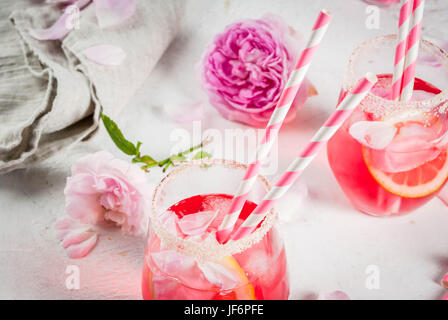 Sommer-Erfrischung Getränke. Leichte rosa rose Cocktail mit Roséwein, Teerose Blütenblätter, Zitrone. Auf einem weißen Stein Beton Tisch. Mit gestreiften rosa Röhrchen Stockfoto