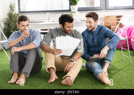 Lächelnde barfuß Jungunternehmer mit Papieren sitzen und mit digital-Tablette, Teamarbeit Geschäftskonzept Stockfoto