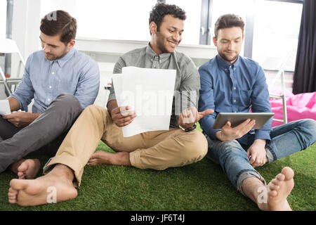 Lächelnde barfuß Jungunternehmer mit Papieren sitzen und mit digital-Tablette, Teamarbeit Geschäftskonzept Stockfoto