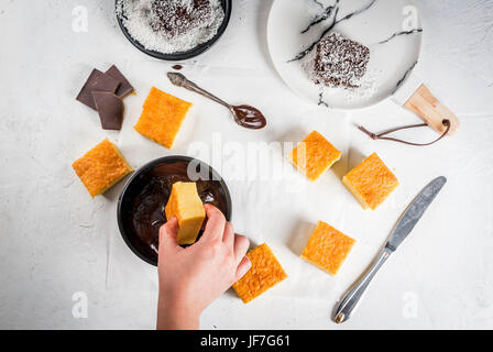 Australische Küche. Vorbereitung australische Dessert Lamington: Biskuit in Schokolade mit Kokos Pulver. Draufsicht, weibliche Hände im Rahmen. Auf ein Jota Stockfoto