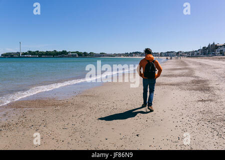 Junger Mann in rote Jacke und Jeans zu Fuß am Strand von Weymouth, Dorset, England an einem klaren sonnigen Tag. Stockfoto