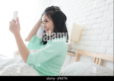 Junge attraktive Asiatin tragen grüne lässige Tücher Selfie Aufnahme beim Sitzen auf dem Bett. Faule Tätigkeit am Sonntagmorgen. Stockfoto