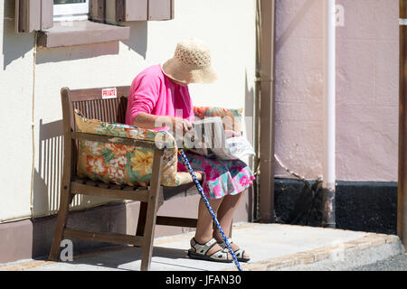 Ältere Frau sitzen, lesen eine Zeitung auf einer Holzbank an der beliebten Uferpromenade Dorf am Kingsand, Cornwall im Sommersonne Sonnenhut tragen und mit einem Gehstock Stockfoto