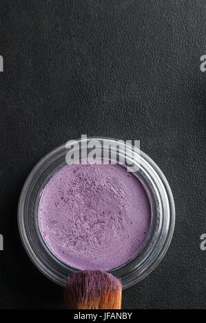 Overhead schließen sich (Makro) ein geöffneten Topf mit violetten Lidschatten, mit Make up Pinsel in Pulver auf Hintergrund schwarz Kunstleder überzogen. Stockfoto