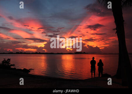 Silhouette paar von Palme beobachten, eine spektakuläre rote, gelbe und lila Sonnenuntergang explodieren hinter dunklen Wolken, alles spiegelt sich in der rote Bucht. Stockfoto