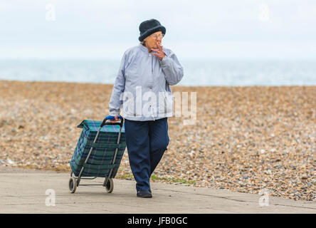 Ältere Dame, die beim Rauchen einen Einkaufswagen zieht, in Großbritannien. Stockfoto