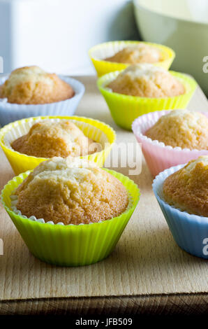 Reihen von Cupcakes, frisch aus dem Ofen, in bunten Silikon Backen Tassen auf einem Holzbrett mit Rührschüssel im Hintergrund. Stockfoto