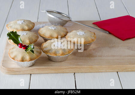 Mince Pies auf einem Holzbrett mit Sternformen und Holly geschmückt.  Eine rote Serviette, alten Teesieb (Sieb) und verstreute Puderzucker ruhen auf den Eber Stockfoto