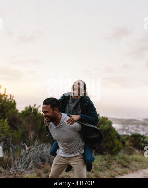 Lächelnder junger Mann mit seiner Freundin auf Rücken. Paar genießt Huckepack Reiten in der Landschaft. Stockfoto