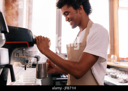 Lächelnd Barista halten Metall Kanne Erwärmung der Milch mit der Kaffeemaschine. Glücklich Jüngling Kaffeezubereitung am Schalter. Stockfoto