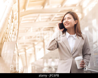 Intelligente asiatische Geschäftsfrau in einem Anzug mit Handy und halten einen Pappbecher Kaffee außerhalb Unternehmenszentrale. Stockfoto