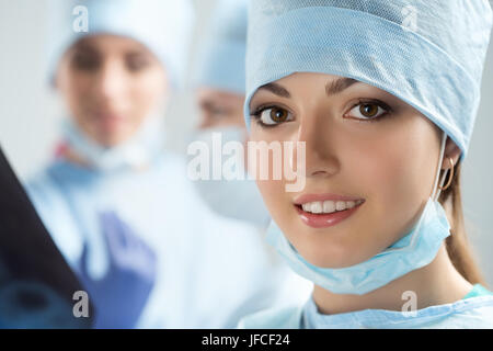 Porträt von glücklichen jungen weiblichen Chirurgen oder Intern nach erfolgreicher Operation mit ihren Kollegen auf Hintergrund im Operationssaal tätig. Healthca Stockfoto