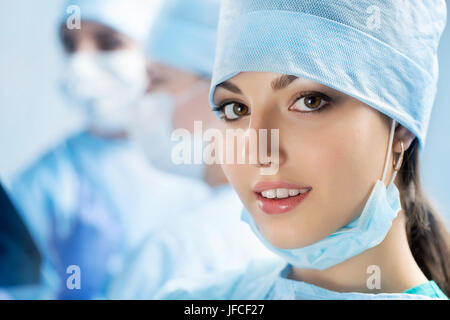 Porträt von glücklichen jungen weiblichen Chirurgen oder Intern nach erfolgreicher Operation mit ihren Kollegen auf Hintergrund im Operationssaal tätig. Healthca Stockfoto