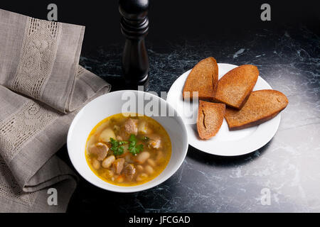 Bohnensuppe in Schüssel auf einem schwarzen Stein Hintergrund weiß. Einige toast auf weißen Teller und schwarze Mühle für Pfeffer auf braunem Tuch. Stockfoto