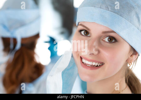 Porträt von glücklichen jungen weiblichen Chirurgen oder Intern nach erfolgreicher Operation mit ihren Kollegen auf Hintergrund im Operationssaal tätig. Healthca Stockfoto