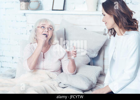 Die Pflege der alten Generation. Lächelnde qualifizierte junge Krankenschwester im Schlafzimmer sitzen und Beratung ältere Frau während ihrer Patientin, die Einnahme von Pillen Stockfoto