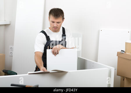 Frustriert Mann gekleidet in Arbeitnehmer insgesamt Anleitung lesen und Selbstmontage Möbel zusammenstellen. DIY, Heim- und bewegliche Konzept Stockfoto